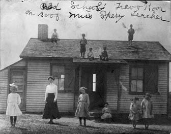 vintge photo of children standing in front of, and on the roof of, old schoolhouse